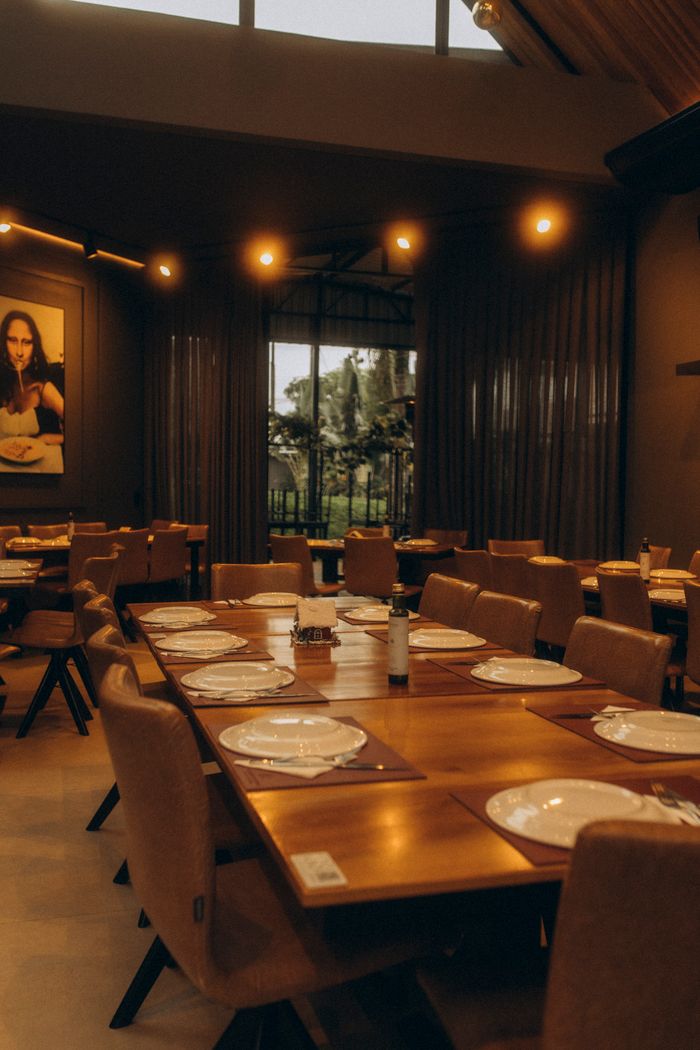 Warmly lit restaurant interior with empty wooden dining tables set with plates and cutlery.